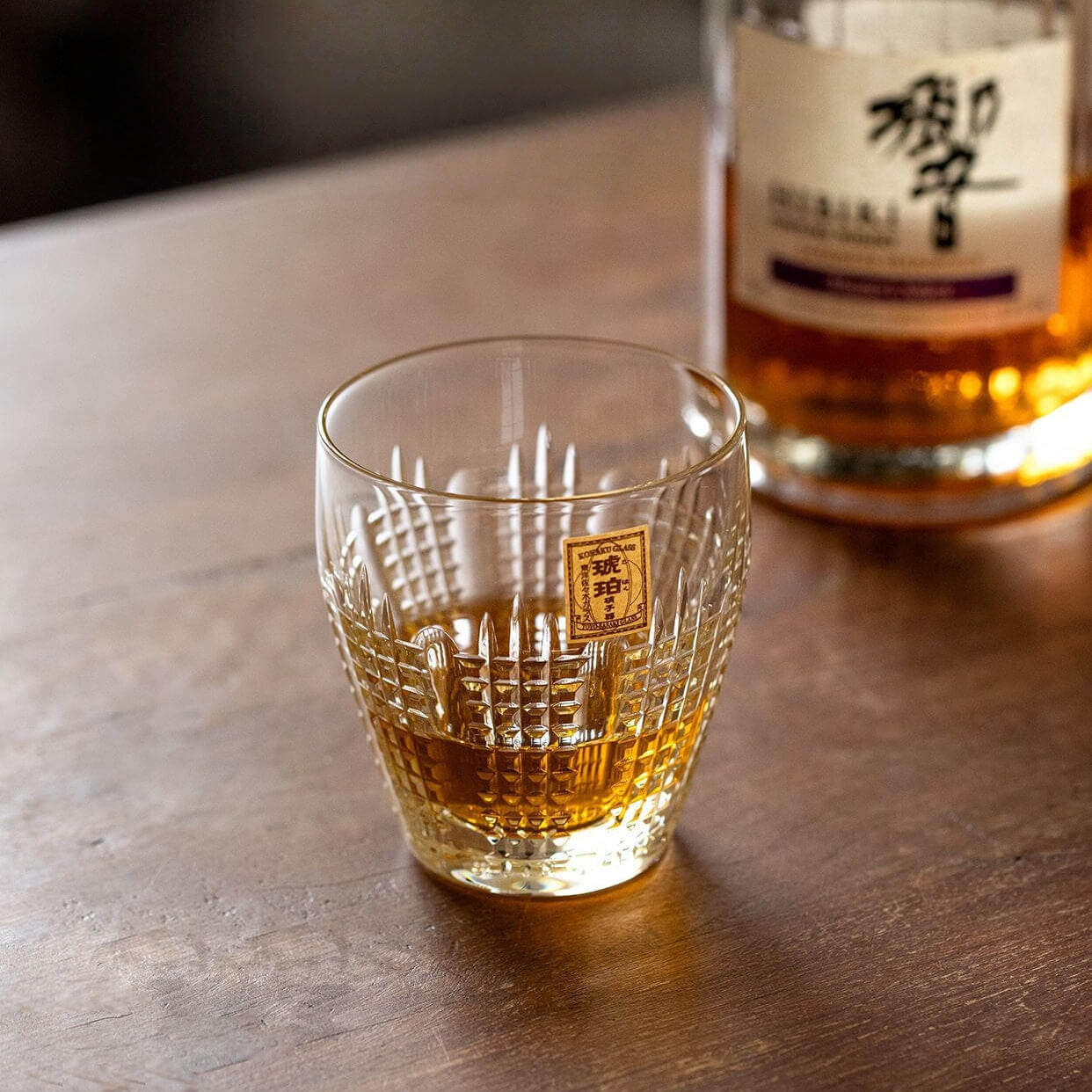 Amber drink in a Kohaku Nanako glass on a wood table with a whiskey bottle behind it